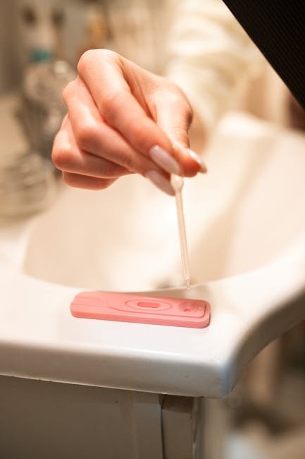 A hand using a pipette on a pregnancy test kit in a bathroom setting.